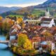 Vista aérea de cidade suíça com rio, ponte, casas coloridas, igreja e colinas arborizadas no outono.