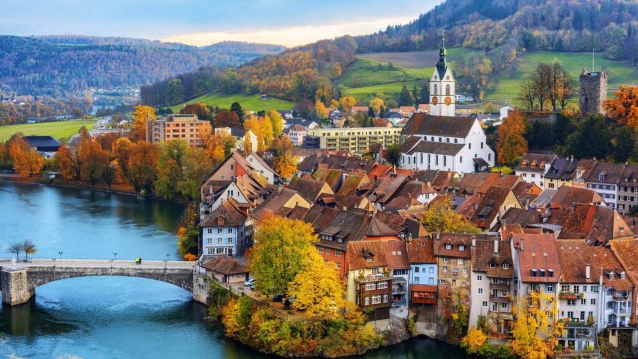 Vista aérea de cidade suíça com rio, ponte, casas coloridas, igreja e colinas arborizadas no outono.