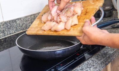 Mãos colocam frango temperado de tábua em frigideira preta sobre fogão de indução ligado.