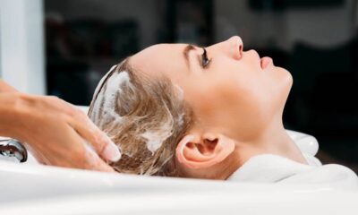 Mulher loira em salão, cabelo sendo lavado com shampoo por mãos de profissional.