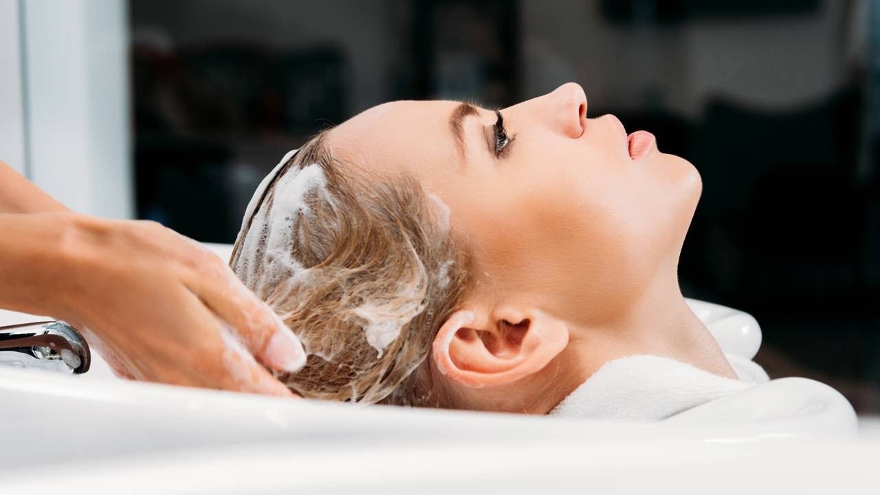 Mulher loira em salão, cabelo sendo lavado com shampoo por mãos de profissional.