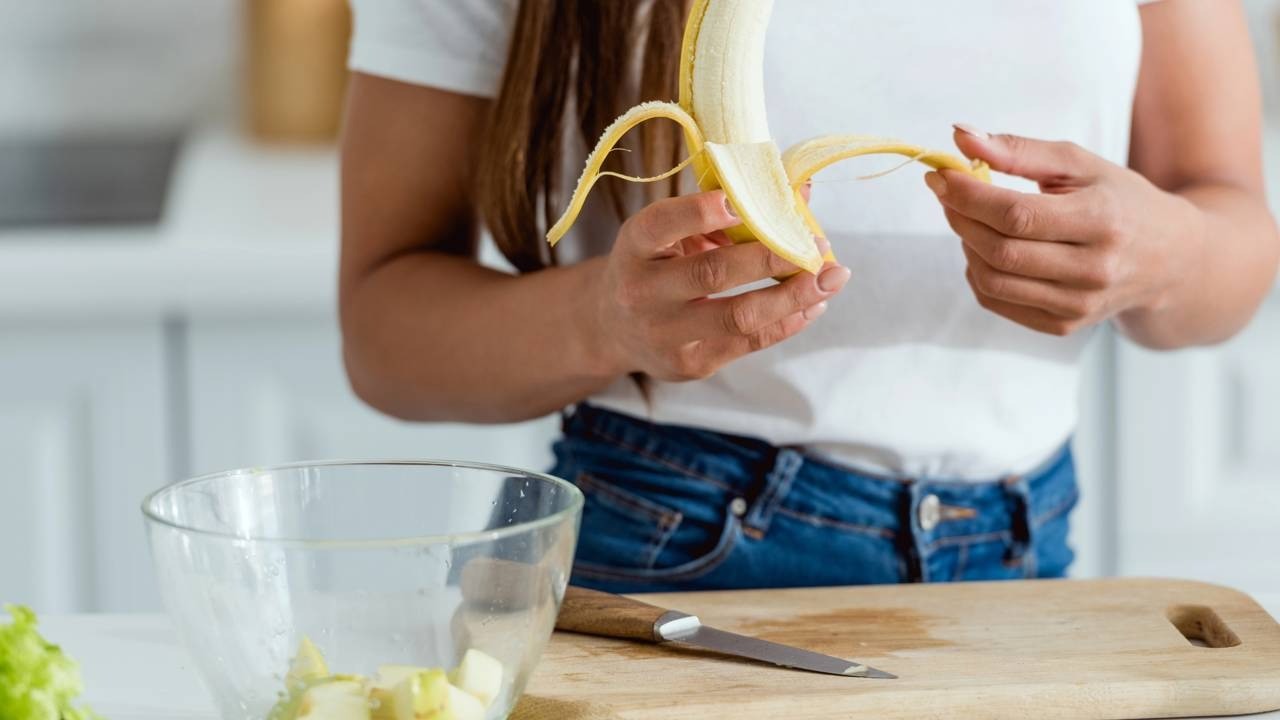 Mãos descascando uma banana sobre uma tábua de corte com uma tigela.
