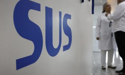 O logo azul 'SUS' em destaque na parede branca, com duas pessoas desfocadas ao fundo.