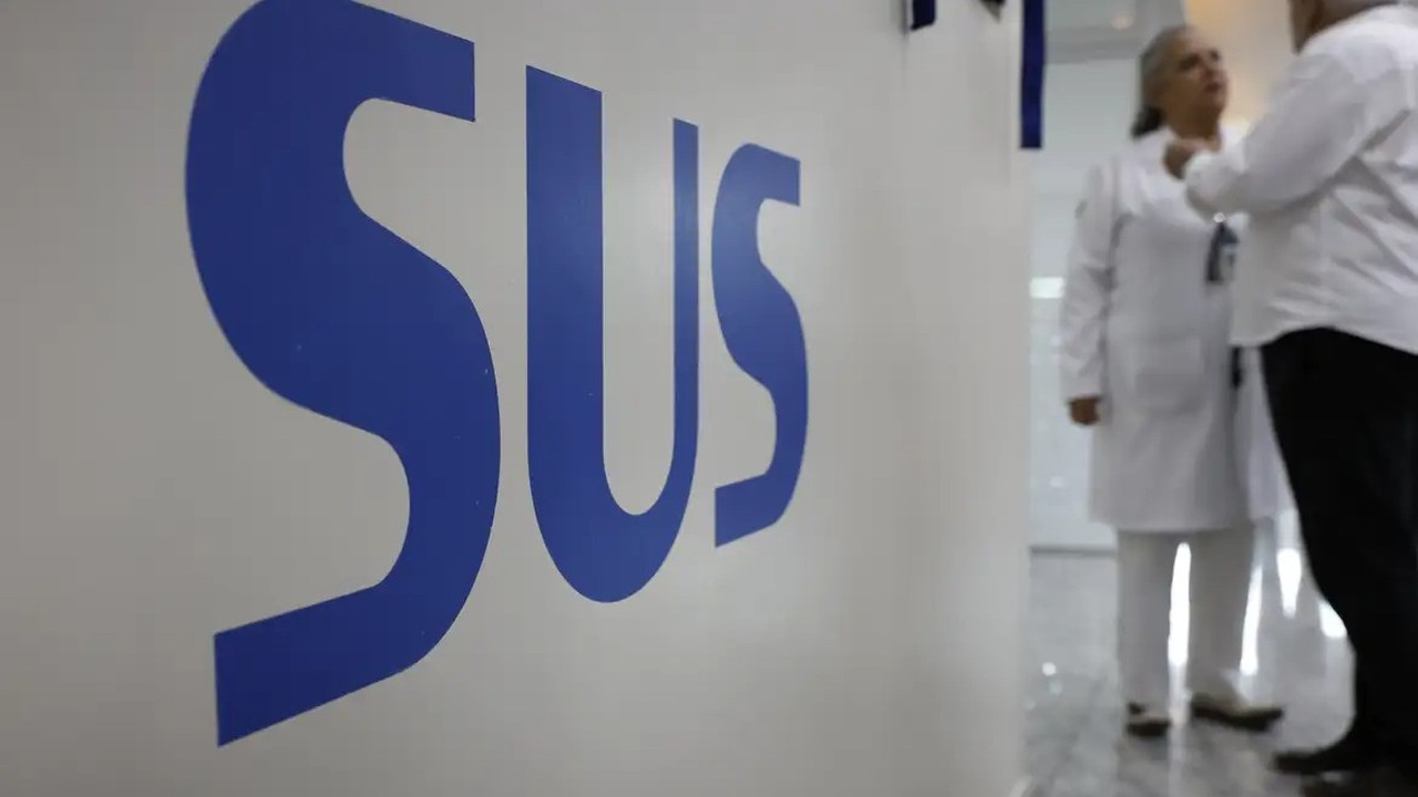 O logo azul 'SUS' em destaque na parede branca, com duas pessoas desfocadas ao fundo.