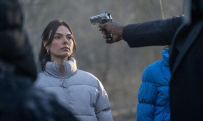 Mulher em jaqueta cinza-clara com arma prateada apontada para sua cabeça.