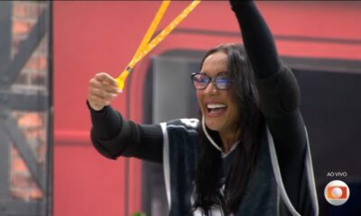 Mulher de óculos, Maxiane Rodrigues, sorrindo e segurando objetos amarelos brilhantes em celebração.