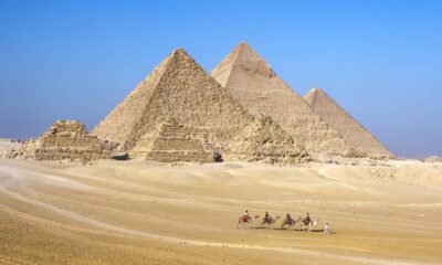 Três pirâmides egípcias no deserto de Gizé com pessoas montadas em camelos no primeiro plano.
