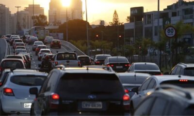 Trânsito congestionado de carros e moto em rua urbana com prédios e pôr do sol.