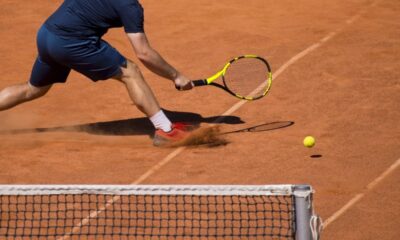 Jogador de tênis deslizando em quadra de saibro, rebatendo bola com raquete amarela e preta.