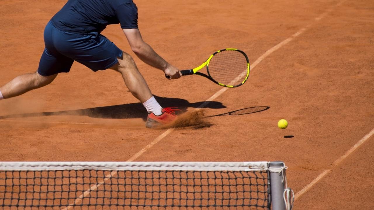Jogador de tênis deslizando em quadra de saibro, rebatendo bola com raquete amarela e preta.