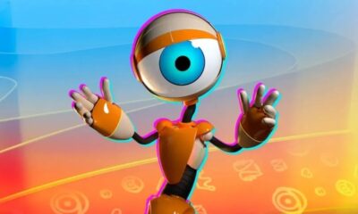 Robô laranja e branco com olho azul gigante, mãos erguidas em fundo colorido.