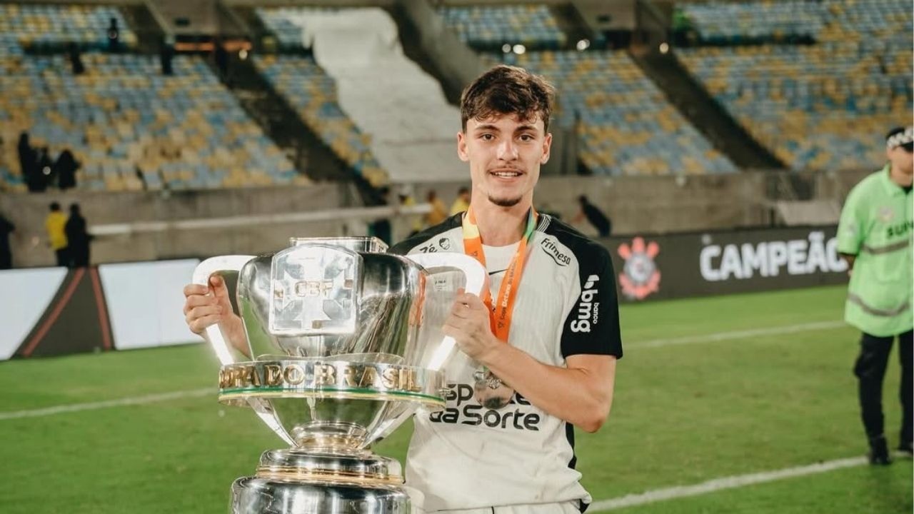 Jovem jogador sorridente com camisa preta e branca segura troféu Copa do Brasil em estádio vazio.