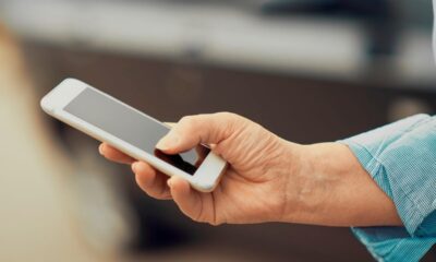 Mão de pessoa adulta segurando um smartphone branco com a tela escura, o polegar toca a tela.