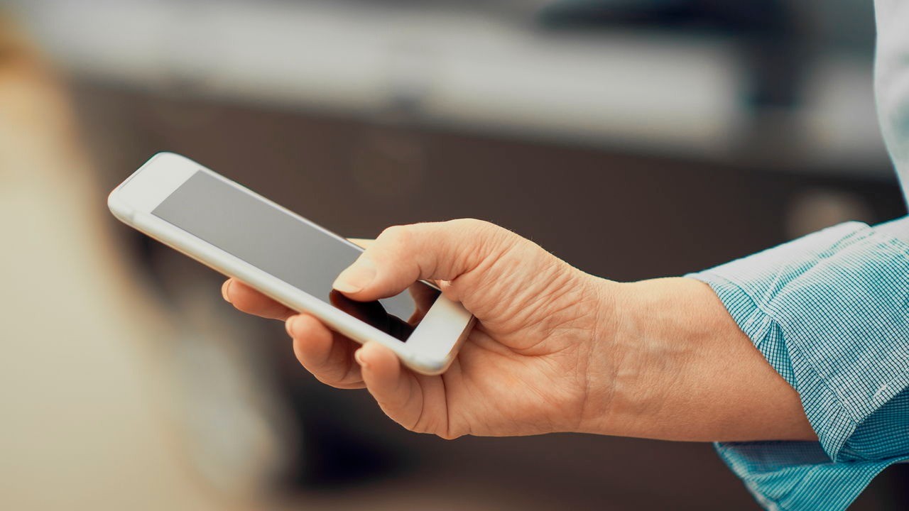Mão de pessoa adulta segurando um smartphone branco com a tela escura, o polegar toca a tela.