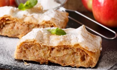Duas porções de strudel de maçã com açúcar de confeiteiro, hortelã, maçãs vermelhas e peneira.