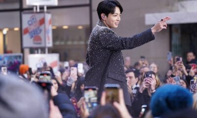 Jungkook do BTS sorrindo em blazer preto brilhante, acenando para fãs com celulares.