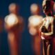 Estatuetas douradas do Oscar, uma em primeiro plano e outras desfocadas ao fundo, contra fundo azul.