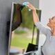 Mulher sorrindo, limpando tela de TV preta montada na parede com pano de microfibra.