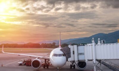 Avião branco em um aeroporto acoplado a um finger, ao pôr do sol, com montanhas.