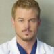 Retrato de Eric Dane, com cabelo loiro, barba, jaleco branco e scrub azul.