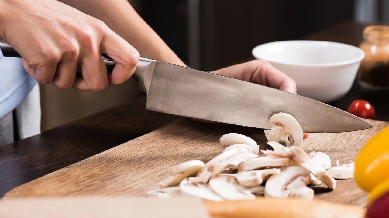 Mãos seguram faca grande fatiando cogumelos brancos em tábua de madeira.