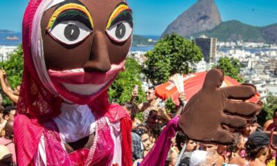 Boneco de Carnaval no meio da multidão, com Pão de Açúcar e cidade do Rio de Janeiro ao fundo.