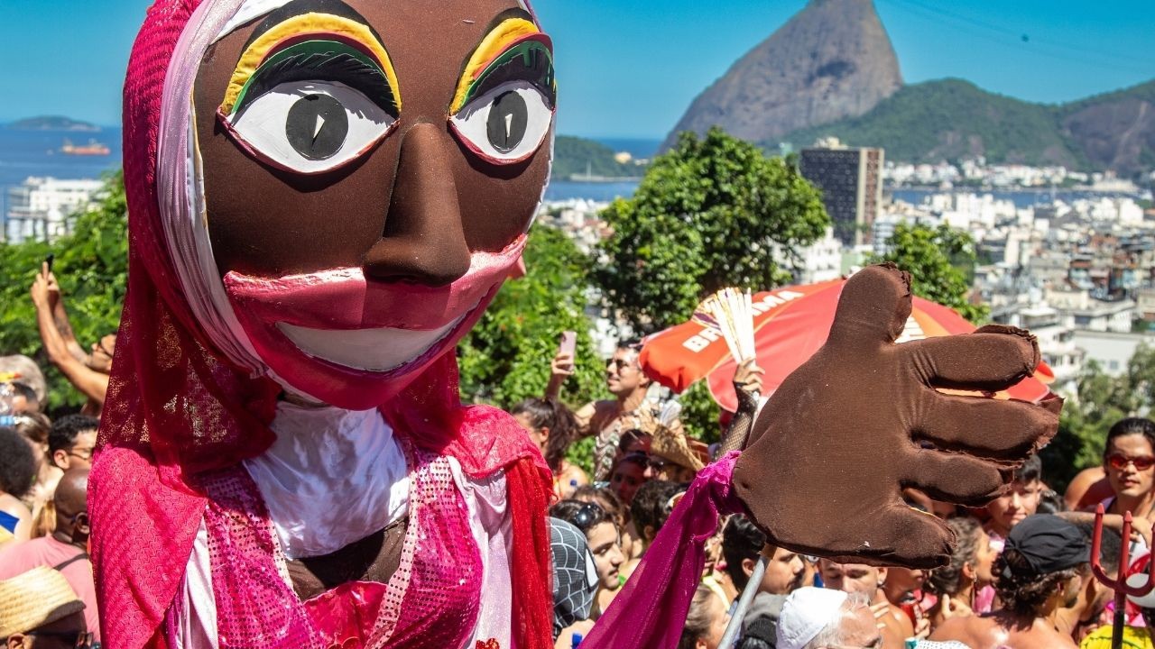 Boneco de Carnaval no meio da multidão, com Pão de Açúcar e cidade do Rio de Janeiro ao fundo.