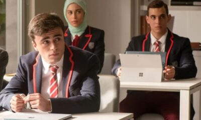 Três jovens em uniformes de colégio: dois homens e uma mulher de hijab em sala de aula.