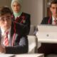 Três jovens em uniformes de colégio: dois homens e uma mulher de hijab em sala de aula.