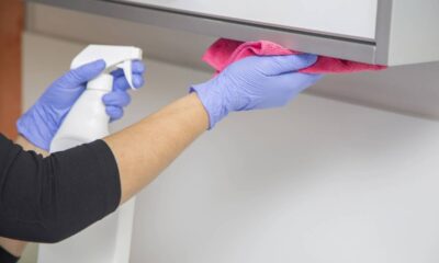 Mãos com luvas azuis limpam armário com spray branco e pano rosa.