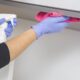 Mãos com luvas azuis limpam armário com spray branco e pano rosa.