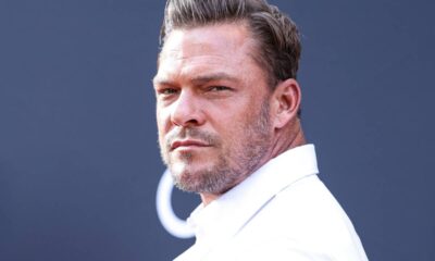 Close-up de Alan Ritchson, homem de cabelo curto grisalho, barba, camisa branca, olhando.