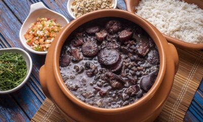 Panela de barro com feijoada, acompanhada de arroz, couve, vinagrete e farofa em mesa rústica.