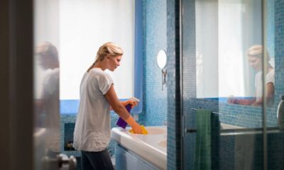 Mulher de cabelo loiro e luvas laranja limpando pia branca de banheiro com spray e esponja.