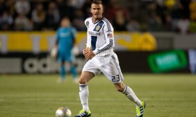 David Beckham com camisa branca e azul, número 23, com bola em campo de futebol.