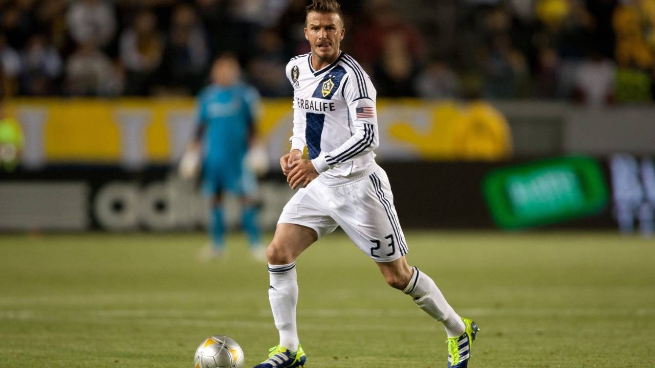David Beckham com camisa branca e azul, número 23, com bola em campo de futebol.