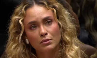 Rosto de Sarah Andrade, cabelo loiro cacheado, olhos claros, brinco, expressão séria.