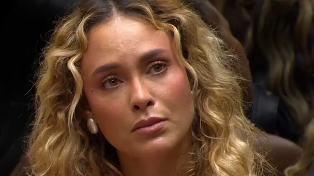 Rosto de Sarah Andrade, cabelo loiro cacheado, olhos claros, brinco, expressão séria.