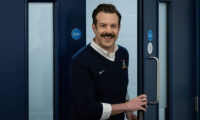 Jason Sudeikis como Ted Lasso, sorrindo e abrindo uma porta em um corredor azul.