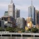 Paisagem urbana de Melbourne com arranha-céus, rio, ponte com bonde e veículos.