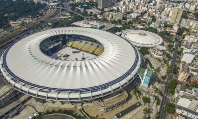 Vista aérea de estádio de futebol circular com teto branco, assentos internos e cidade ao redor.