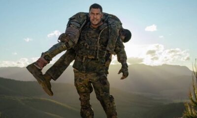 Homem de uniforme militar musculoso carregando outro homem em uniforme, paisagem montanhosa.