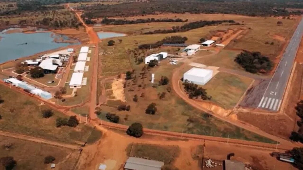 Vista aérea de uma grande fazenda com pista de pouso, edifícios brancos, lago e estradas de terra.