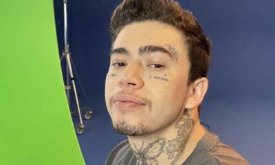 Whindersson Nunes sorrindo levemente, com tatuagens no rosto e pescoço, fundo verde e azul.