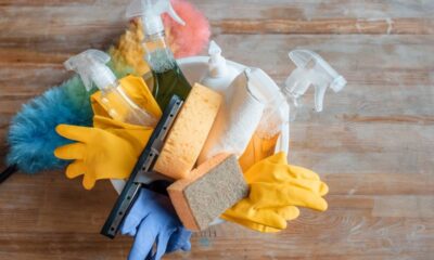 Vários produtos de limpeza, incluindo luvas, borrifadores, esponjas e panos, em um balde branco.