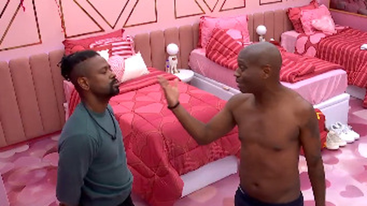 Dois homens em discussão intensa em quarto de reality show, um deles gesticula.