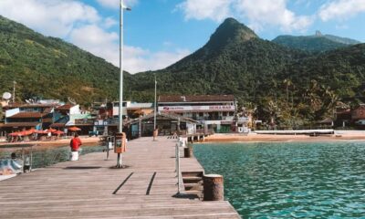 Píer de madeira, pessoa em vermelho, praia com construções, montanhas verdes e mar azul-turquesa.