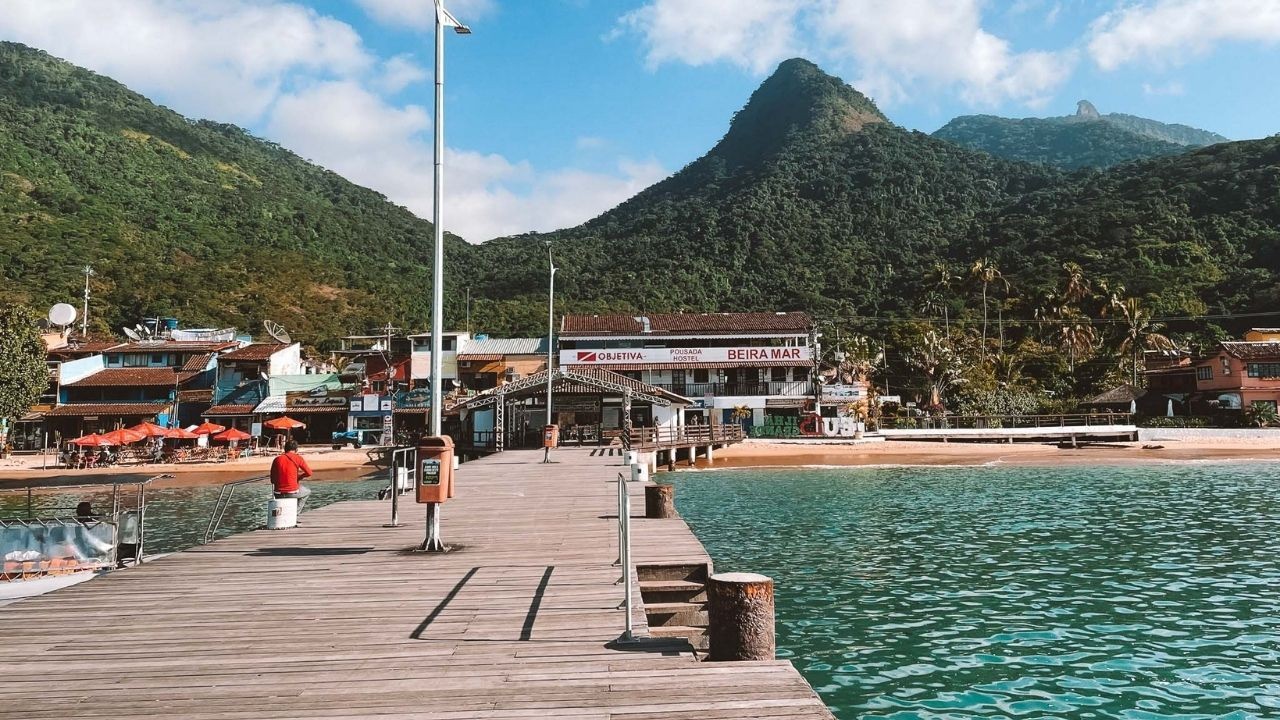 Píer de madeira, pessoa em vermelho, praia com construções, montanhas verdes e mar azul-turquesa.