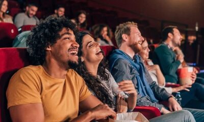 Pessoas diversas riem em sala de cinema, com assentos vermelhos e pipoca.