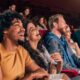 Pessoas diversas riem em sala de cinema, com assentos vermelhos e pipoca.